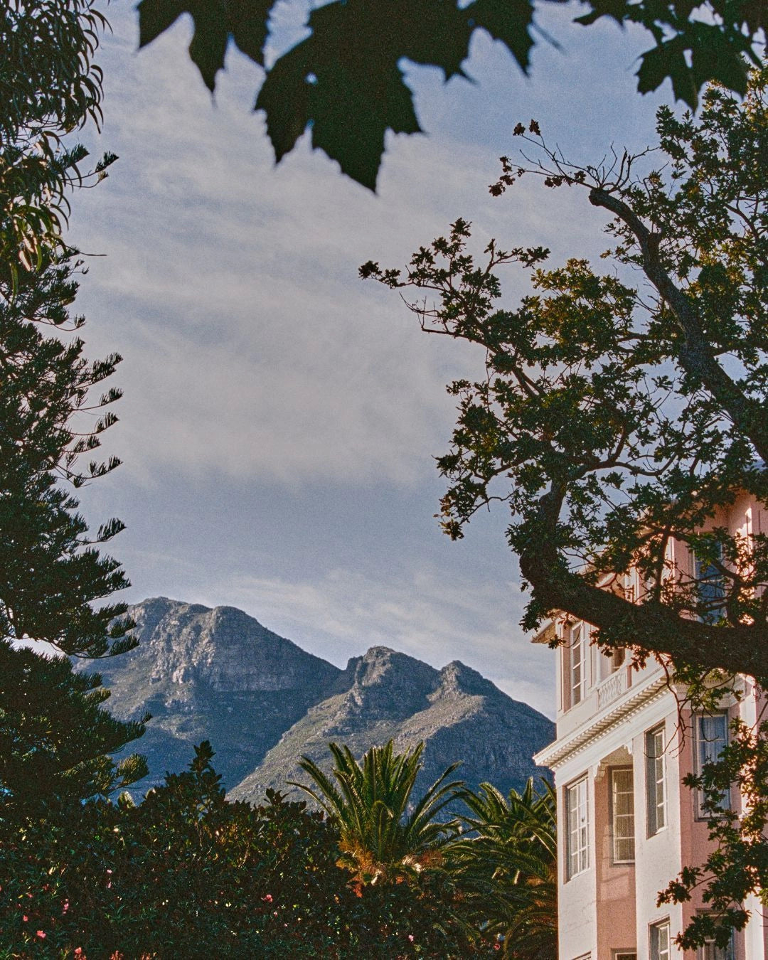 A scenic photograph of a mountain landscape framed by tree branches and leaves, with a pale pink building visible on the right side under a cloudy blue sky.